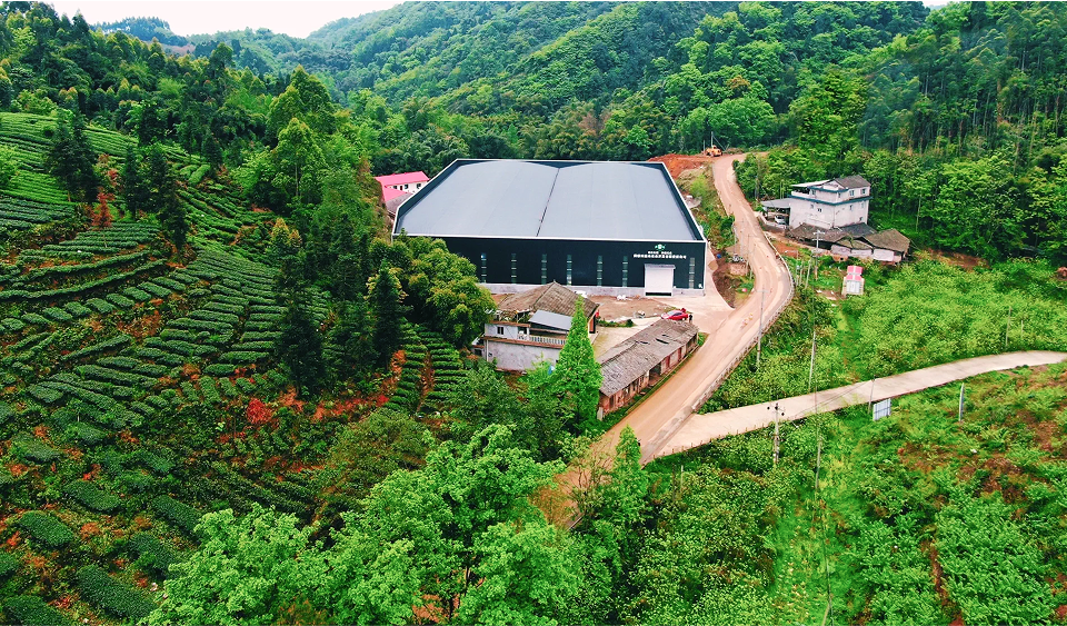 A large industrial building is surrounded by lush green hills, terraced fields, and scattered houses along a winding road.