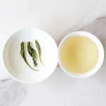 A white bowl with three Biluochun Green Tea leaves from My Store sits next to a cup of pale yellow tea on a white marble surface, rich in antioxidants.