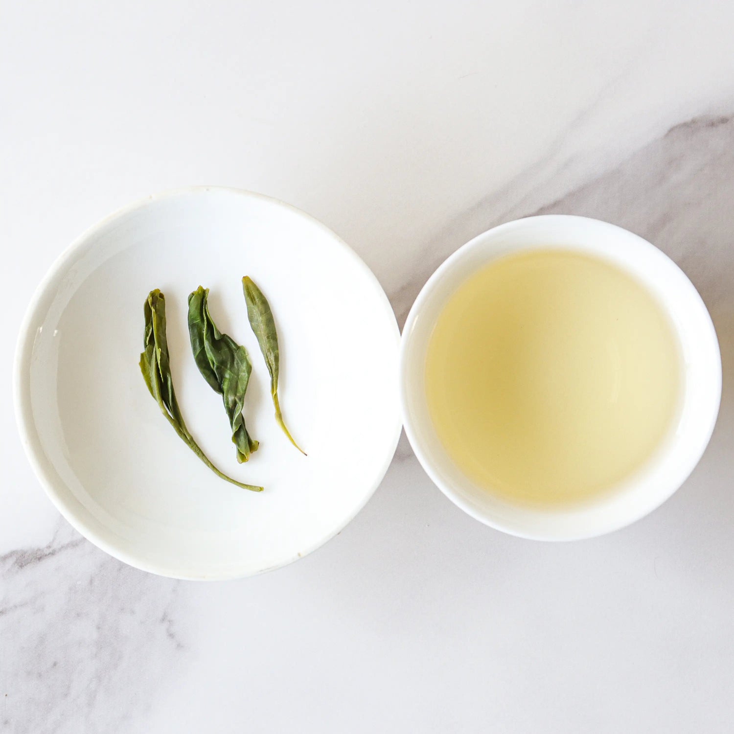 A white bowl with three Biluochun Green Tea leaves from My Store sits next to a cup of pale yellow tea on a white marble surface, rich in antioxidants.