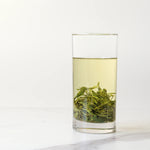 A clear glass filled with light yellow-green Biluochun Green Tea from My Store, with tea leaves settled at the bottom, sits on a white surface against a plain background.