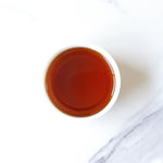 A white cup filled with dark amber Small Chrysanthemum Pu’er Tea Cakes by My Store, viewed from above on a white and light gray marble surface.