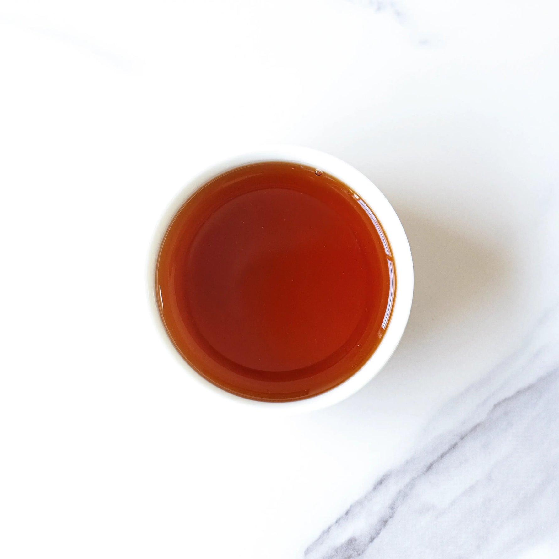 A white cup filled with dark amber Small Chrysanthemum Pu’er Tea Cakes by My Store, viewed from above on a white and light gray marble surface.
