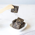 A wooden tong holds a Small Chrysanthemum Pu'er Tea Cake by My Store above a white dish with more tea cakes, all on a white surface.