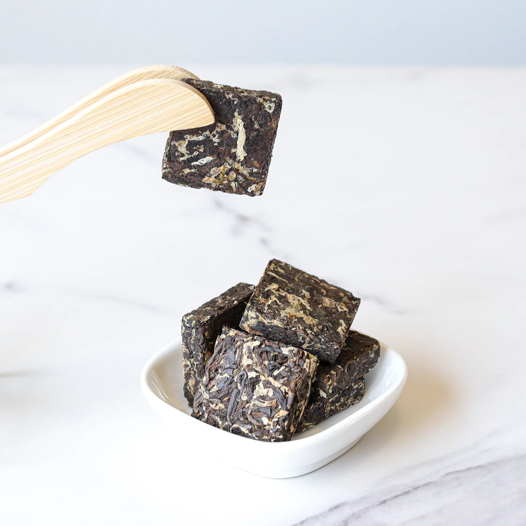 A wooden tong holds a Small Chrysanthemum Pu'er Tea Cake by My Store above a white dish with more tea cakes, all on a white surface.