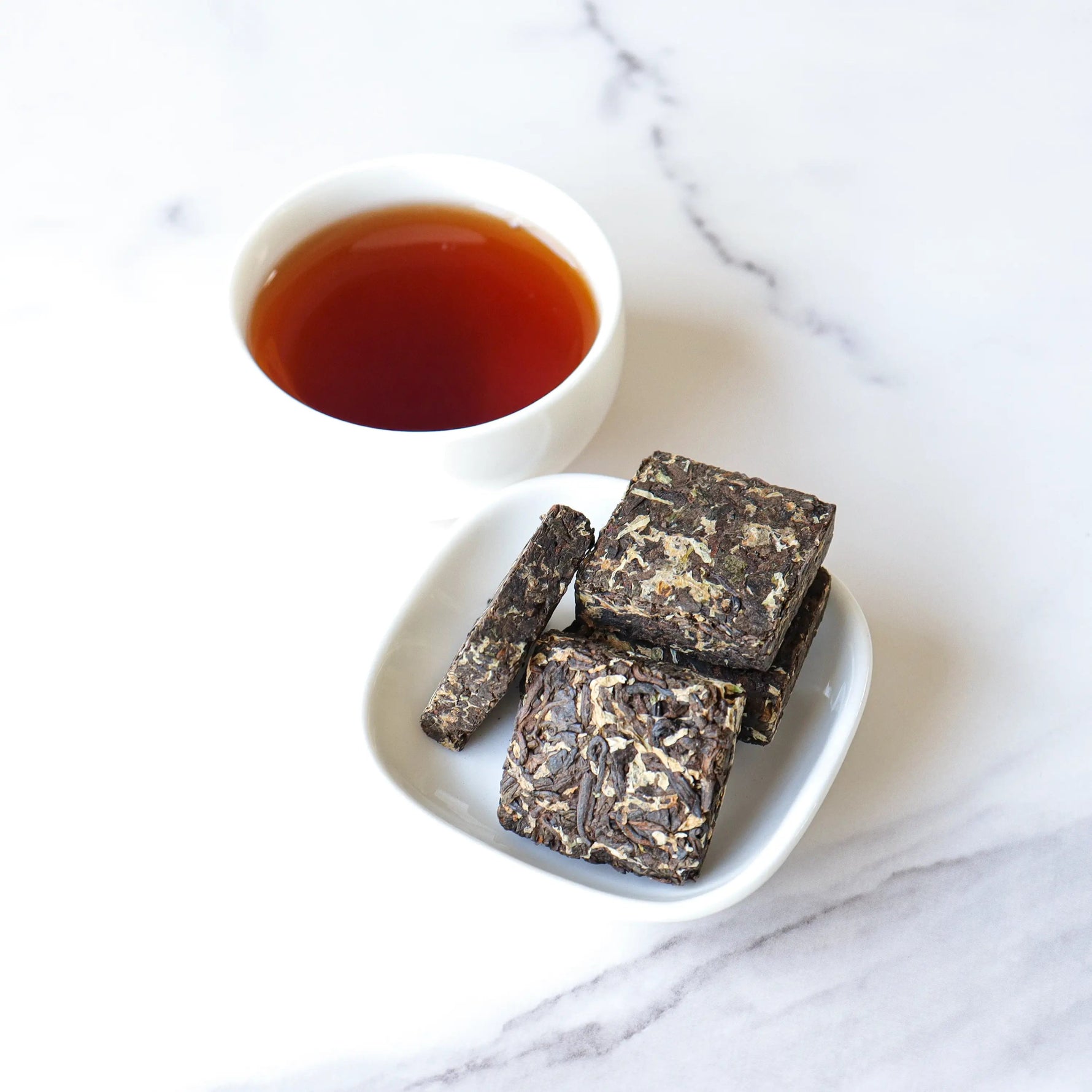 A white cup of tea and a small white plate holding several My Store Small Chrysanthemum Pu’er Tea Cakes sit on a white marble surface.