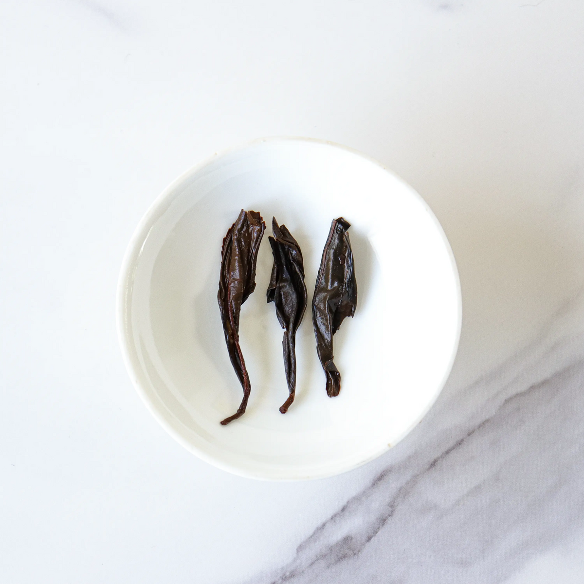 Three Da Hong Pao Oolong Tea leaves in a white bowl on a marble surface.