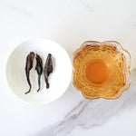 A white dish with three dried Da Hong Pao Oolong Tea leaves from My Store sits beside a glass cup of amber-colored tea on a white marble surface.