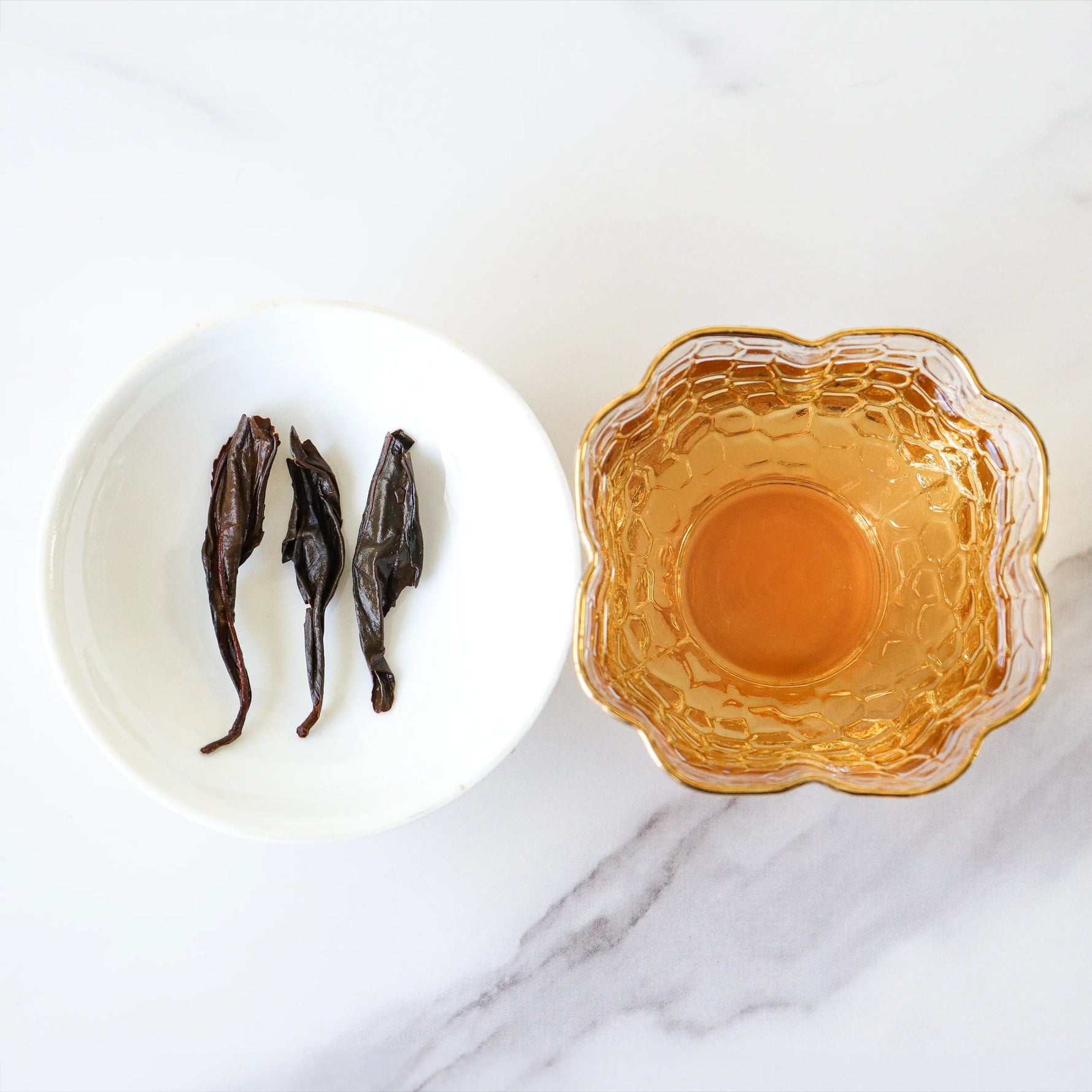 A white dish with three dried Da Hong Pao Oolong Tea leaves from My Store sits beside a glass cup of amber-colored tea on a white marble surface.