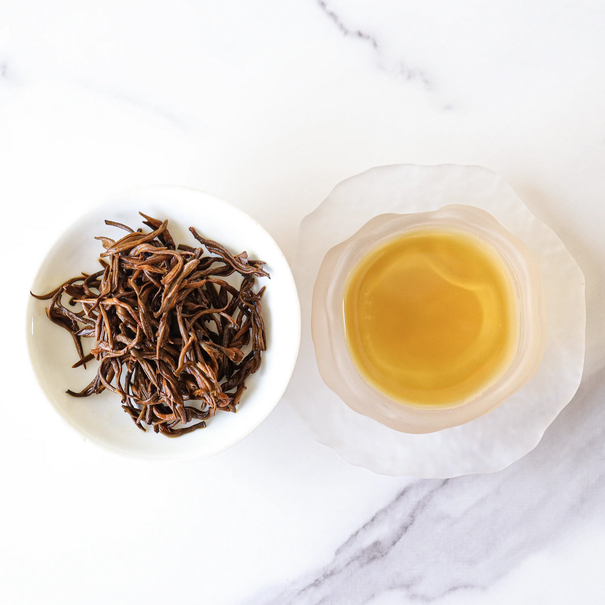 Dianhong black tea leaves beside a clear glass cup of amber tea on marble.