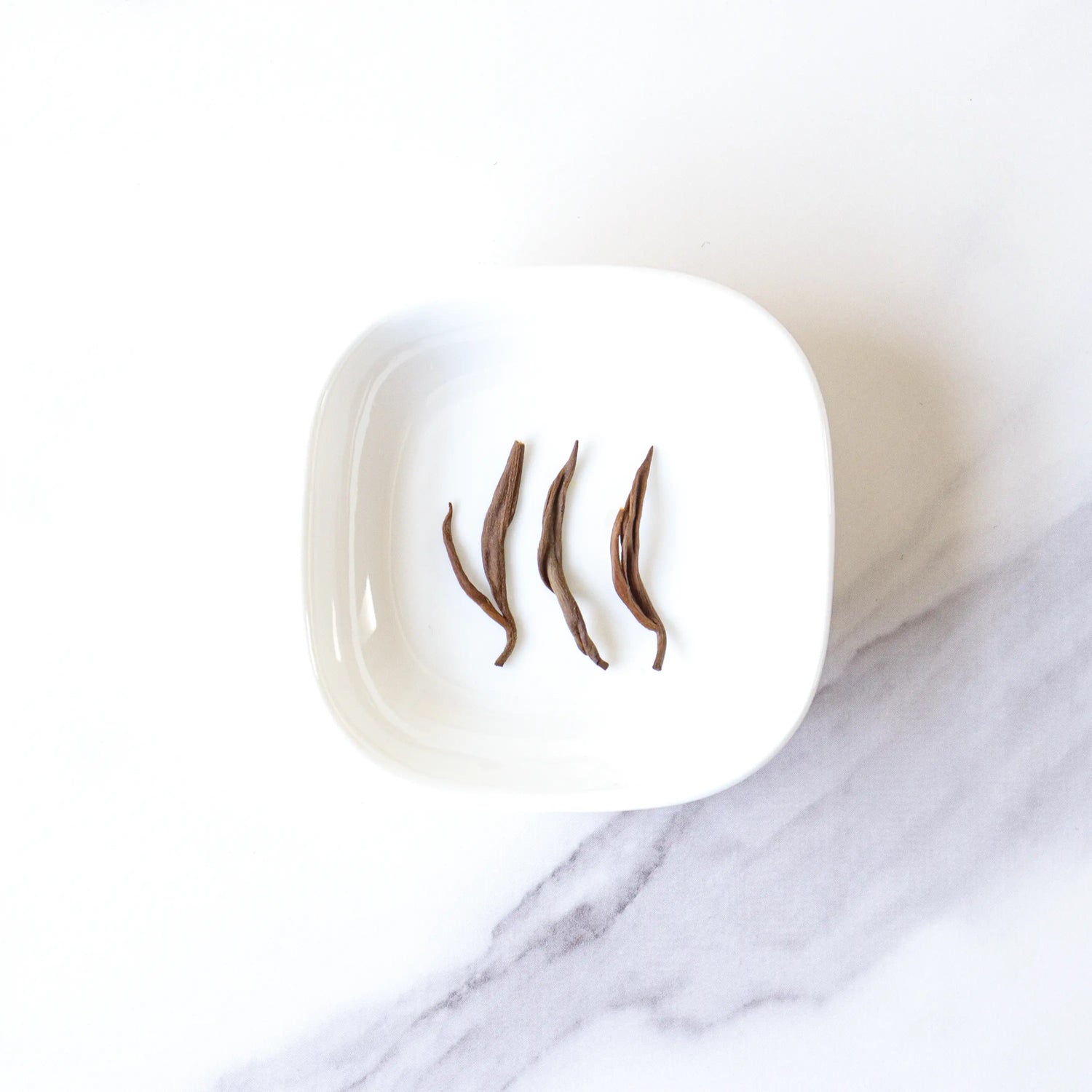 Three dried Dianhong Black Tea leaves from My Store are arranged side by side in a white square dish on a white and gray marbled surface, highlighting this premium black tea.