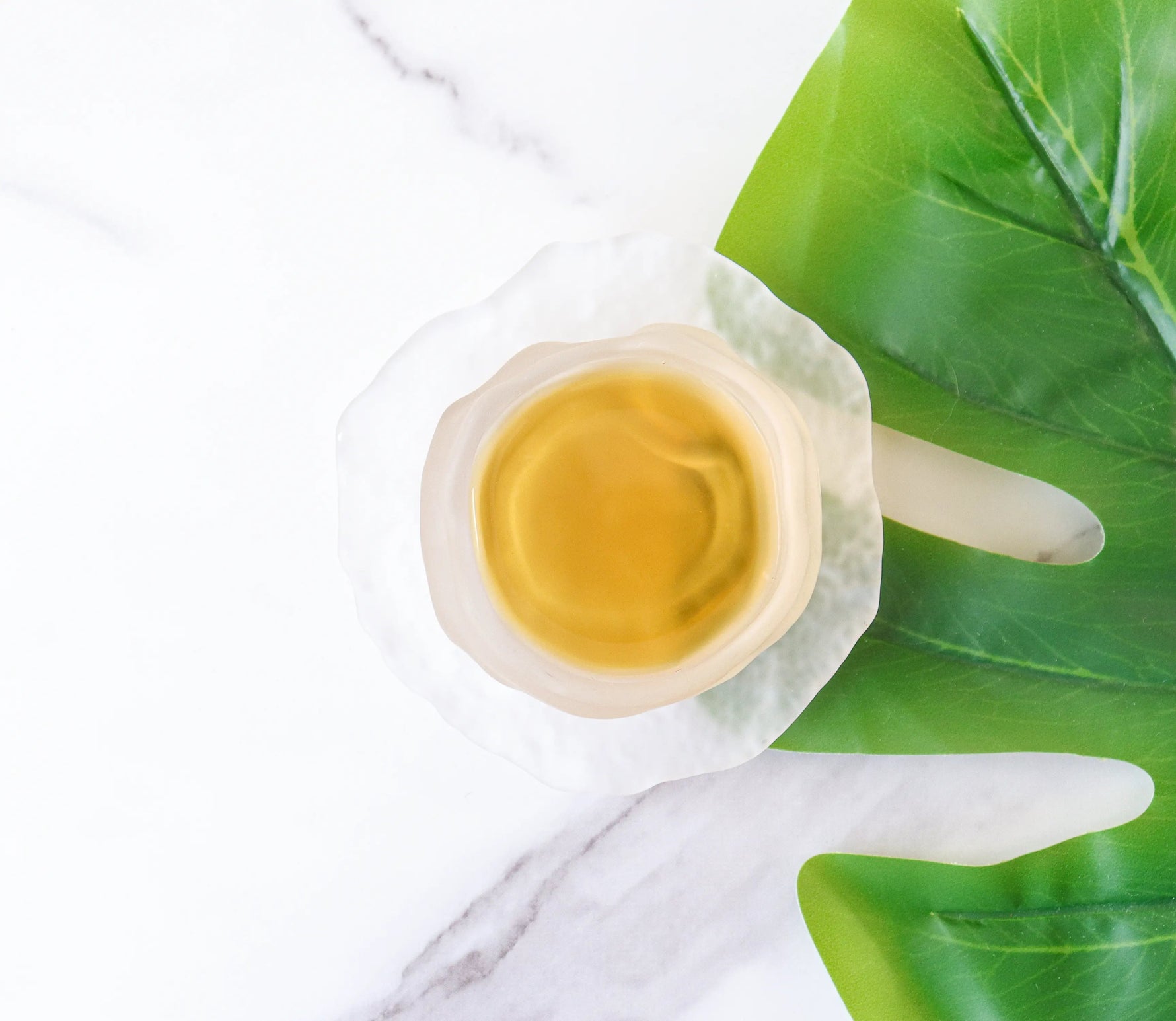 A cup of My Store's Dianhong Black Tea rests on a white marble surface beside large green leaves. This premium tea from high-altitude regions delivers elegant, subtle flavors and features a beautiful light hue.