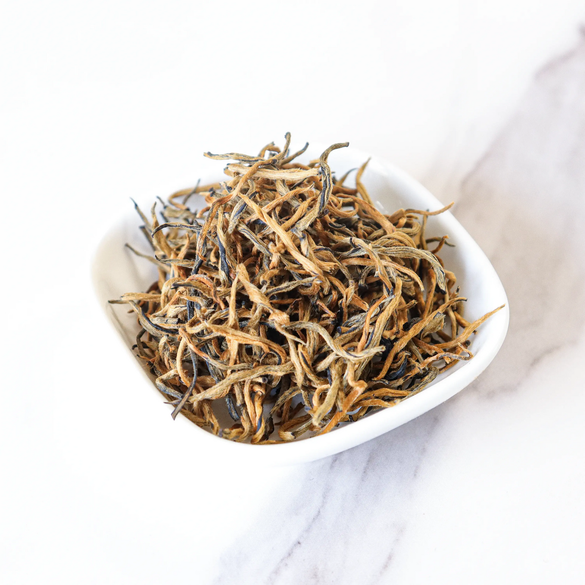 Loose Dianhong black tea leaves with golden tips in a white bowl on marble.