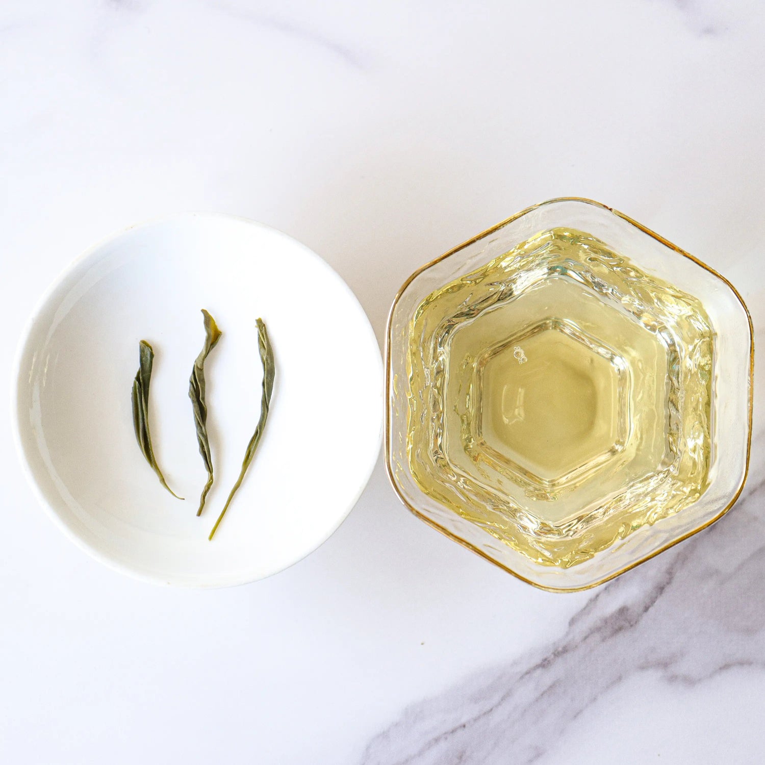 A hexagonal glass cup filled with My Store's Emei Maofeng Green Tea sits beside a small white dish holding three tea leaves on a white marble surface.