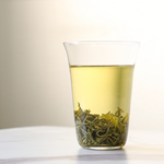 A glass of light yellow-green Emei Maofeng Green Tea from My Store, with loose tea leaves settled at the bottom, sits on a white surface against a plain background.