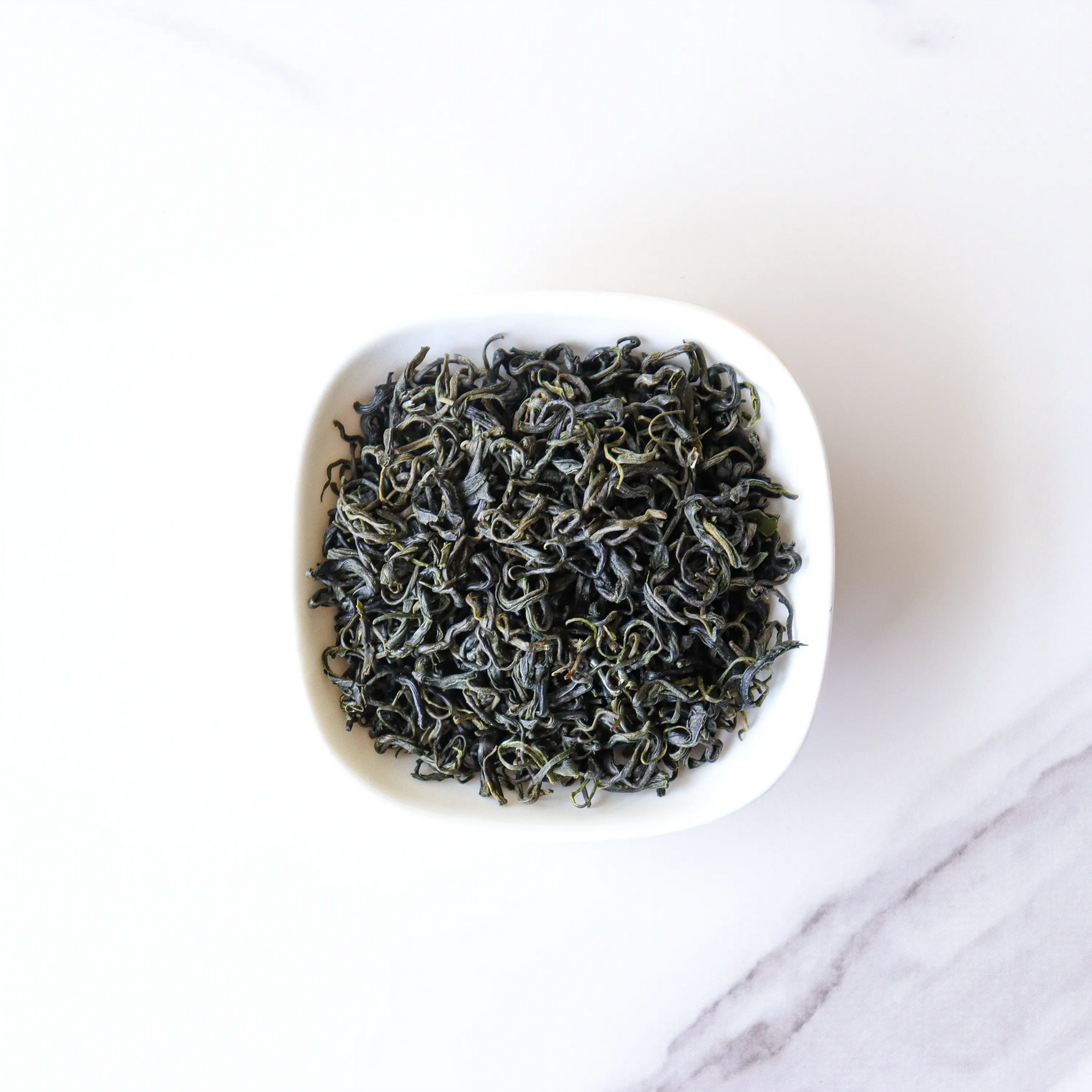A small white bowl holds loose, dried Emei Maofeng Green Tea leaves from My Store, displayed on a white marble surface and highlighting this distinctive tea from Sichuan Province.