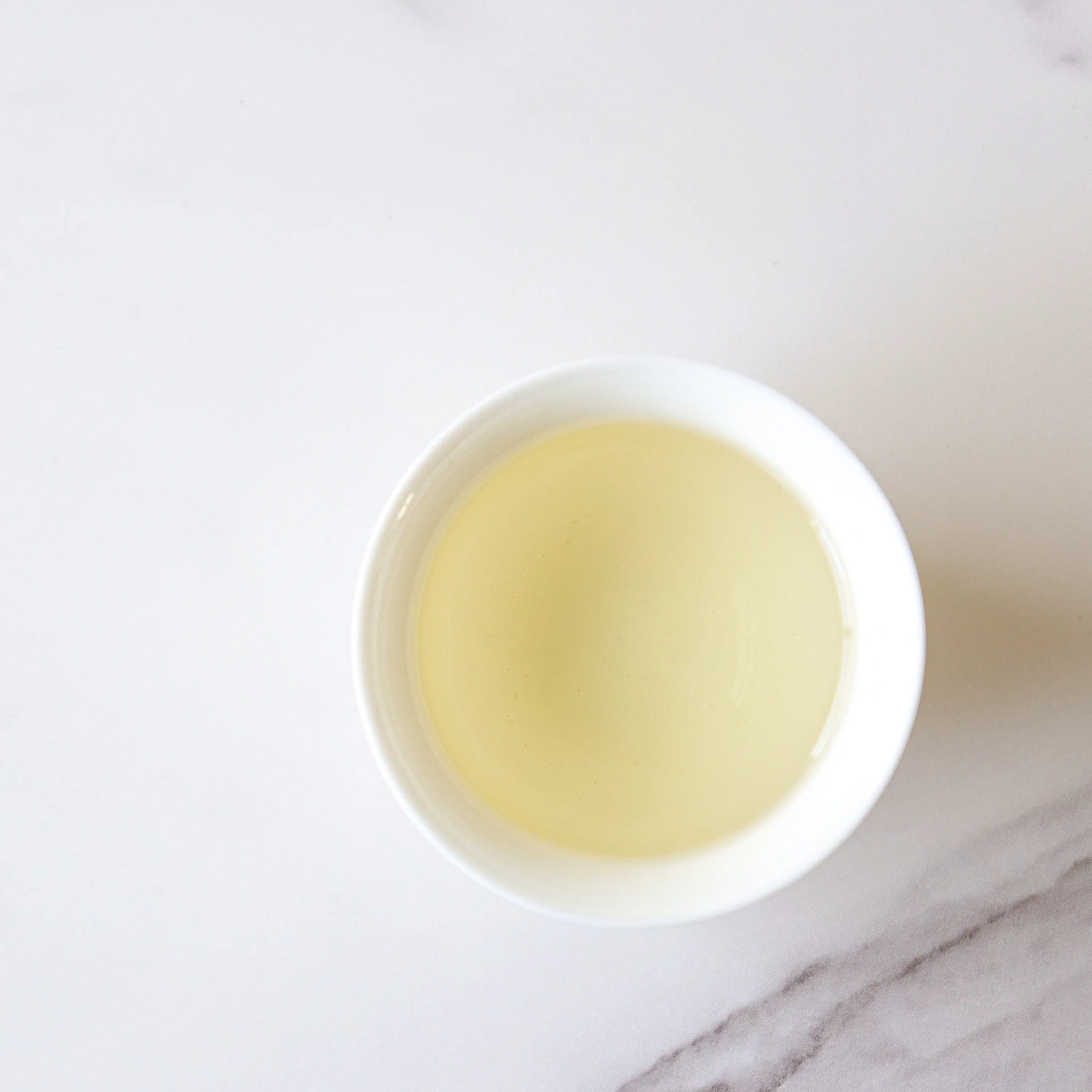 A white cup of light yellow-green Enshi Yulu Green Tea by My Store sits on a white marble surface, capturing the premium loose-leaf tea's essence.