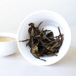 A white bowl tipped on its side holds wet, steeped Gong Mei White Tea leaves from My Store beside a cup of brewed tea, set on a white surface.