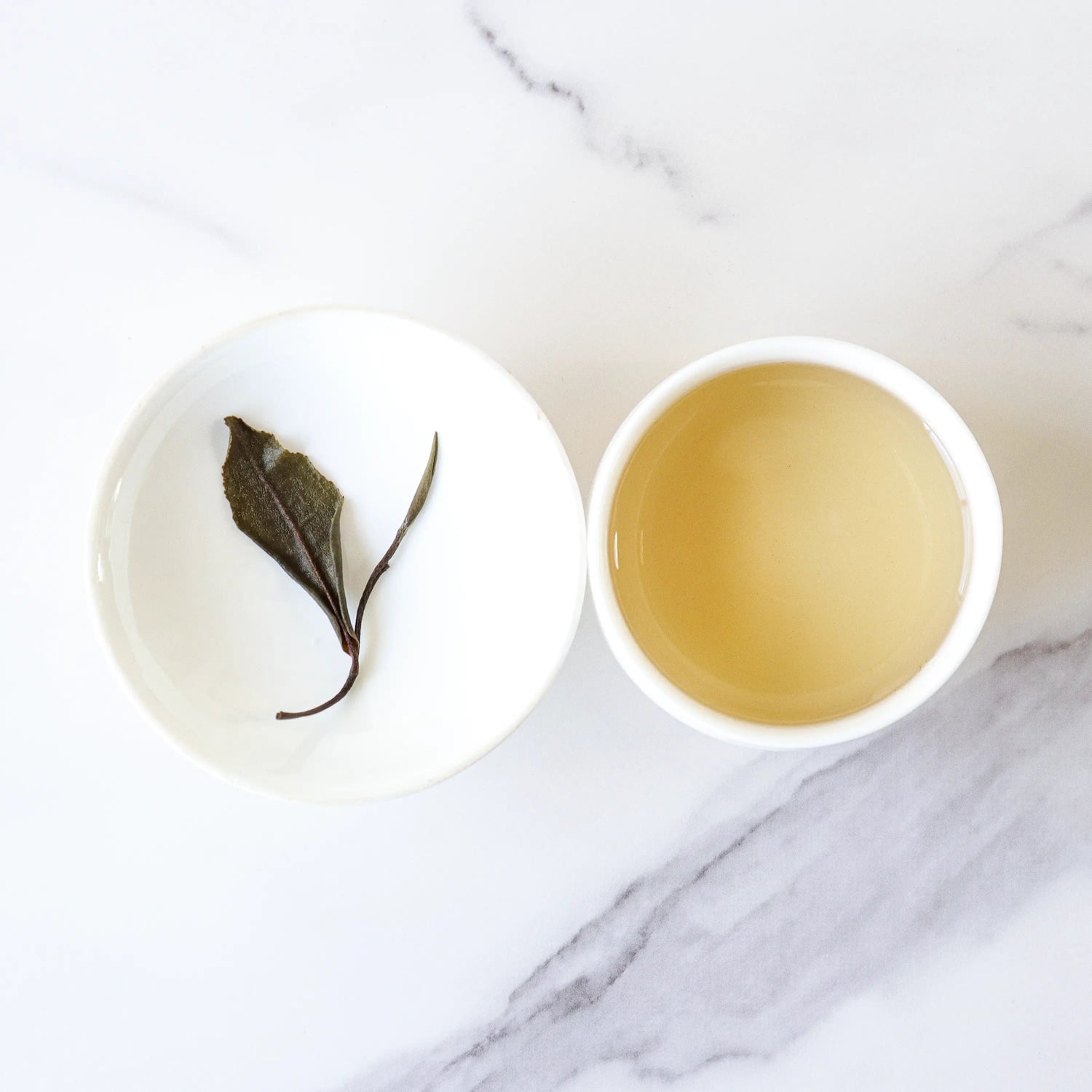A cup of light-colored Gong Mei White Tea by My Store sits beside a small dish with a single steeped leaf on white marble, highlighting the tea’s elegant simplicity from Fuding City.