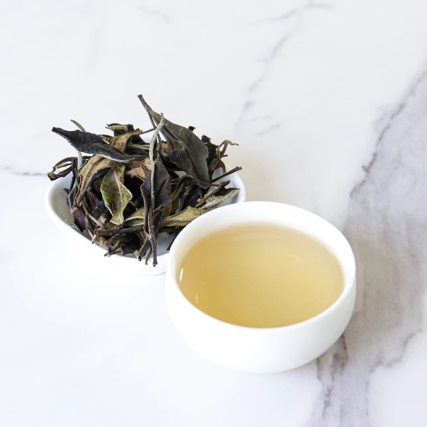 A small white bowl of My Store's Gong Mei White Tea sits beside a matching cup filled with pale brewed tea from Fuding City, all on a white marble surface.