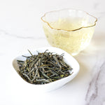 A small bowl of My Store Huangshan Maofeng Green Tea loose leaves sits next to a clear glass cup of brewed tea on a white marble surface.