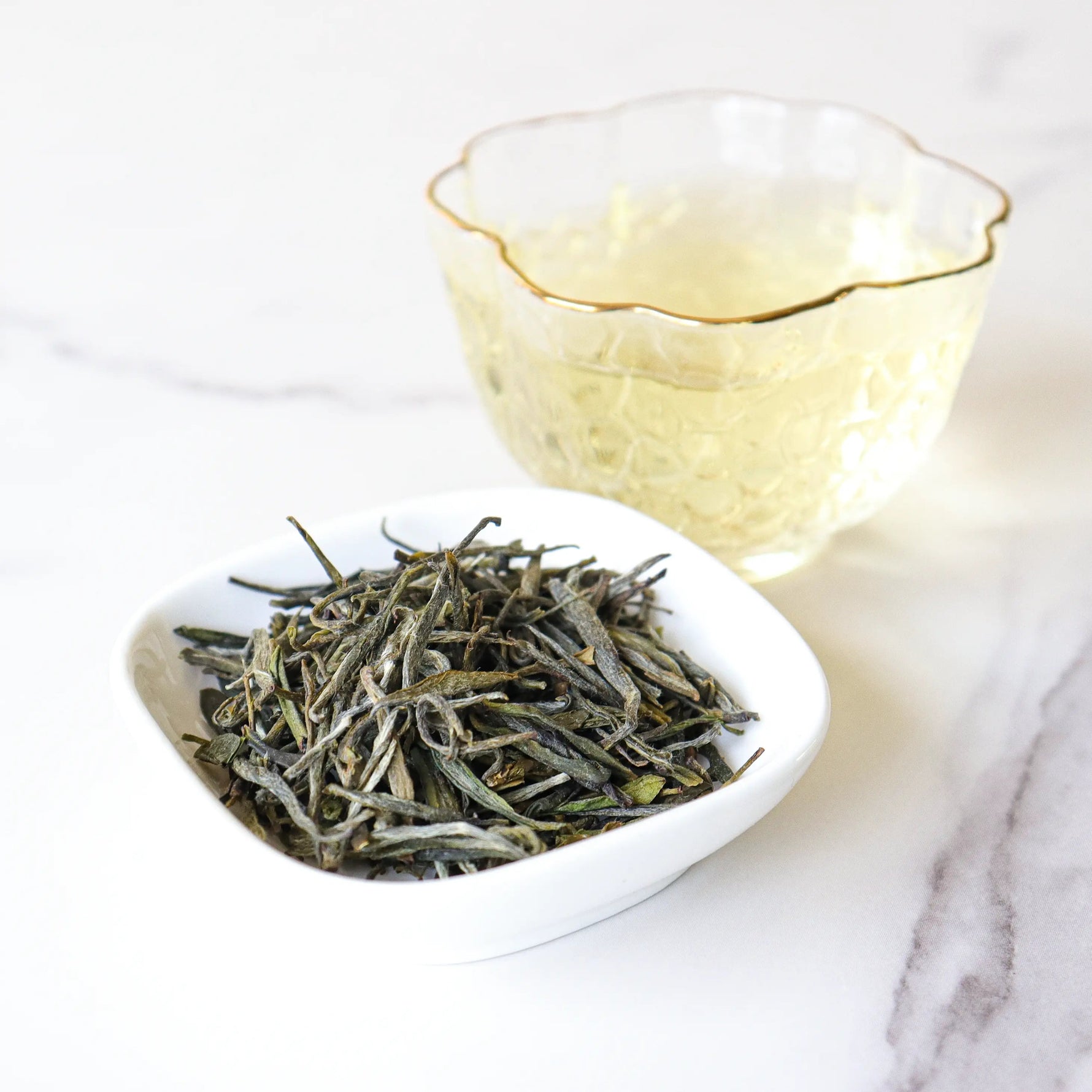 A small bowl of My Store Huangshan Maofeng Green Tea loose leaves sits next to a clear glass cup of brewed tea on a white marble surface.