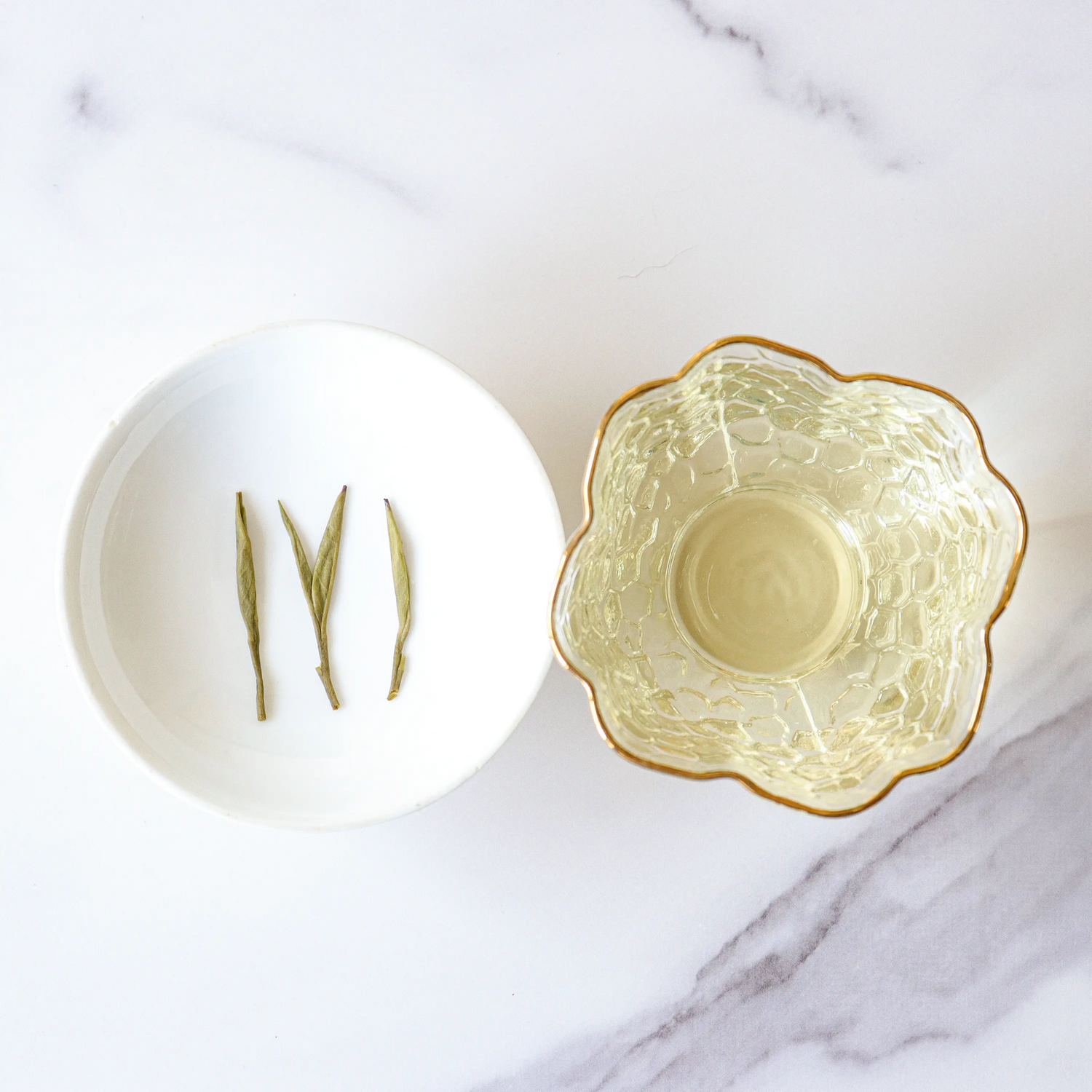 A white dish with four green tea leaves sits beside a clear glass cup of light Huangshan Maofeng Green Tea from My Store, both elegantly arranged on a white marble surface.