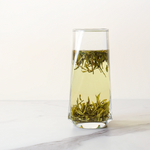 A clear glass of Huangshan Maofeng Green Tea from My Store, with loose tea leaves floating in the light-colored brew, sits on a white surface against a plain background.