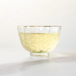 A clear glass bowl with a honeycomb pattern holds light yellow Huangshan Maofeng Green Tea from My Store, set on a white surface against a plain background.