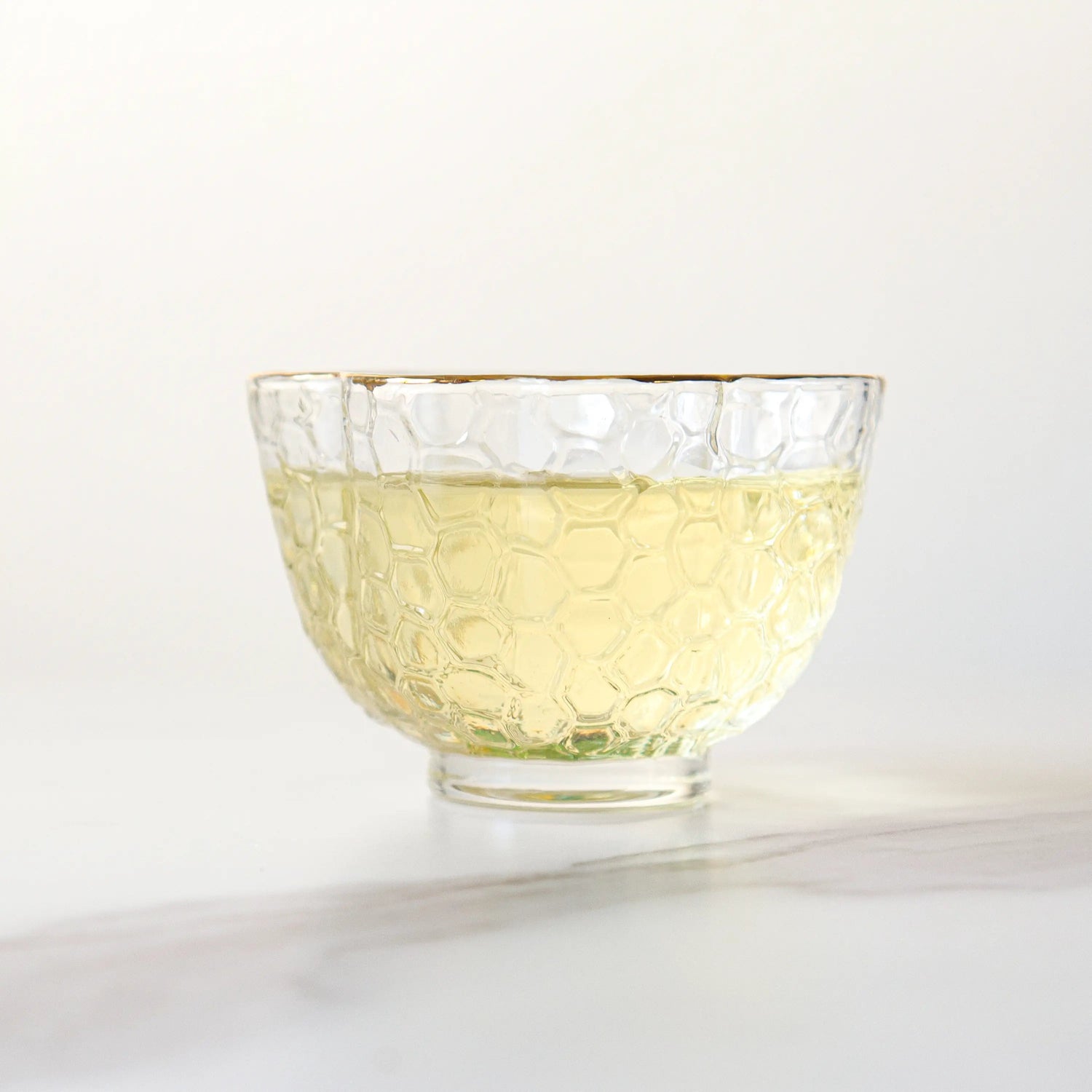 A clear glass bowl with a honeycomb pattern holds light yellow Huangshan Maofeng Green Tea from My Store, set on a white surface against a plain background.