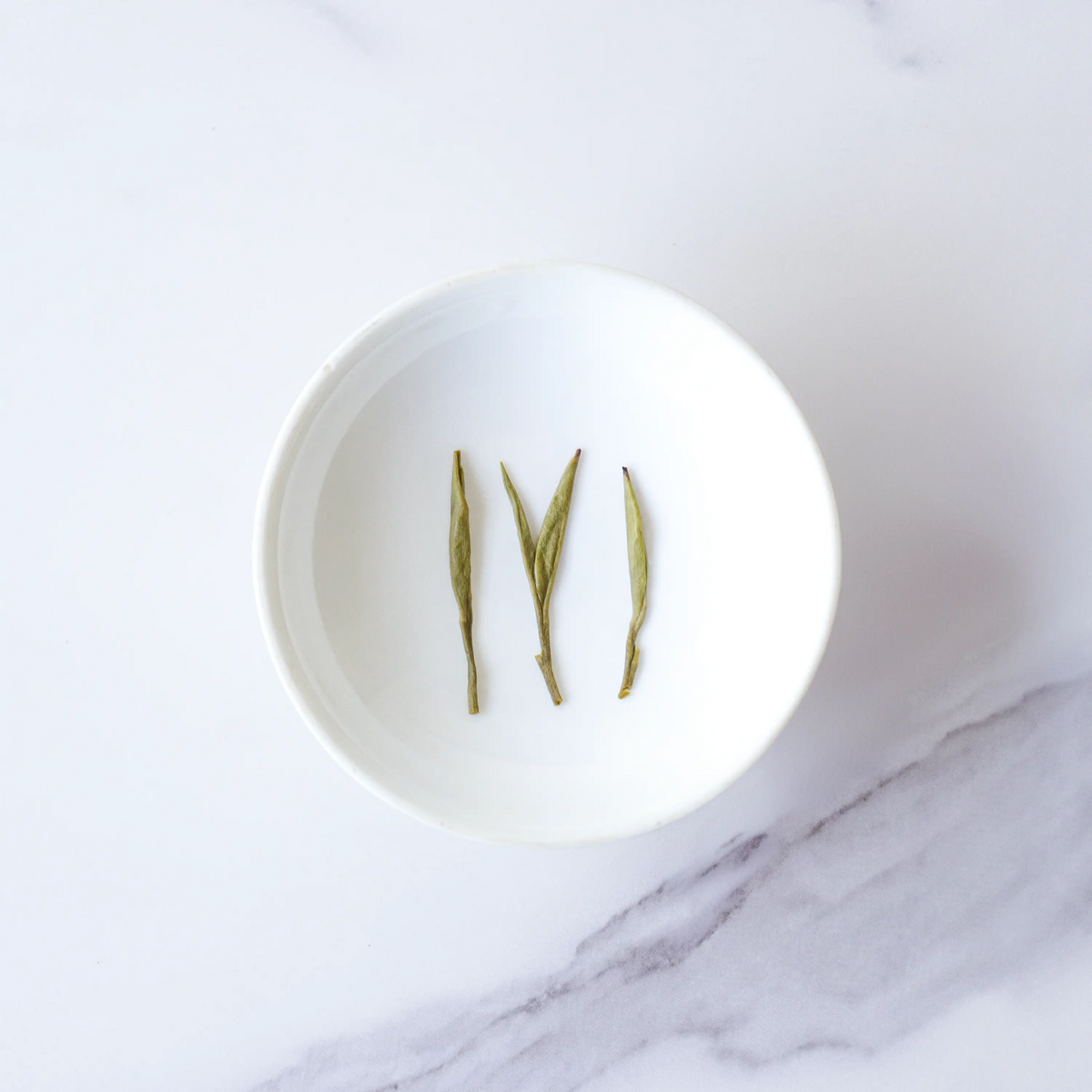 Huangshan Maofeng Green Tea leaves arranged in a white bowl on a marble surface.