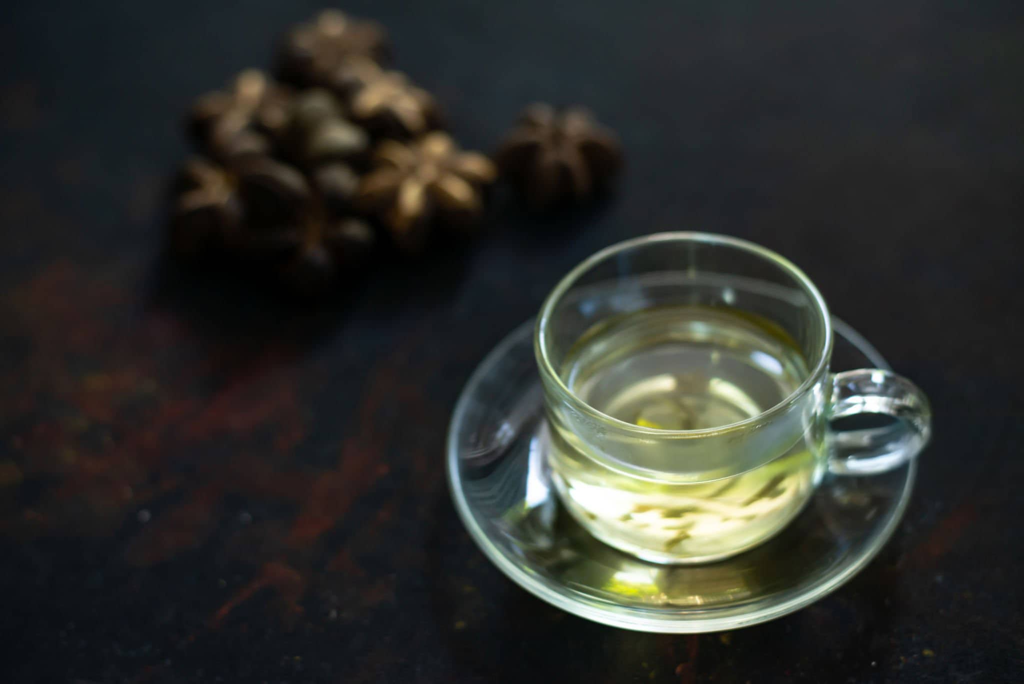 A clear glass cup and saucer with light-colored tea, with star anise pods in the background on a dark surface.