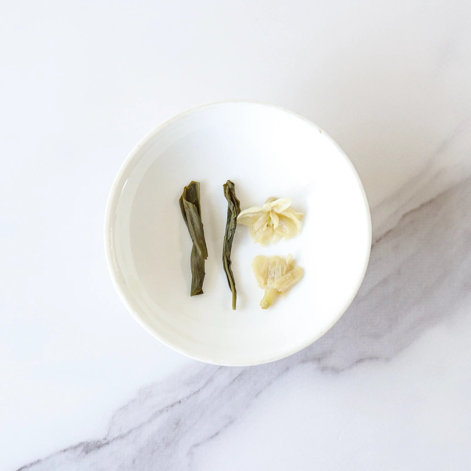 A white bowl with two green tea leaves and two white flower petals, capturing the delicate essence of My Store Jasmine Tea, sits on a light marble surface.