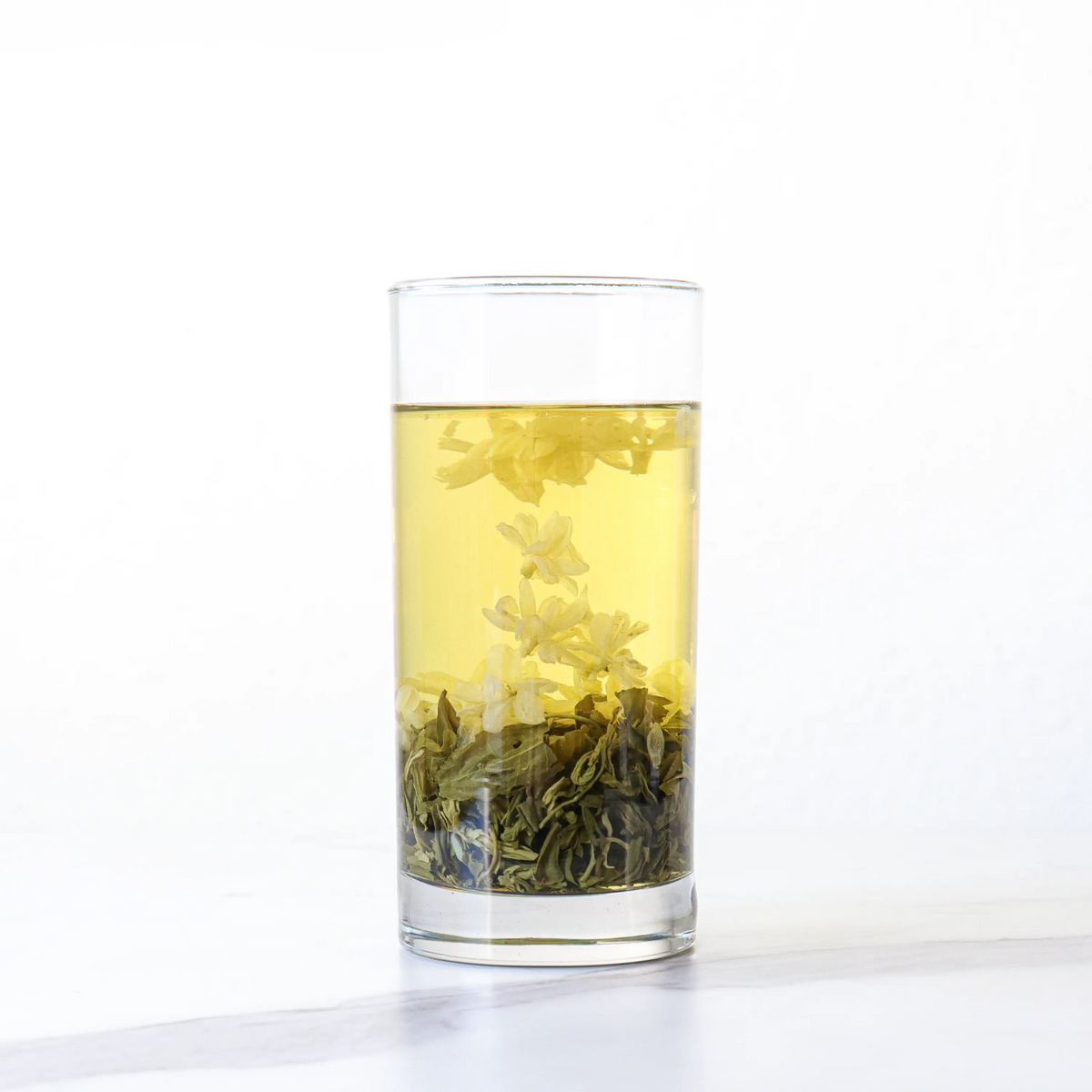 Jasmine Green Tea brewing in a tall glass with jasmine blossoms and loose leaves.