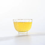A small, clear glass cup with a textured pattern holds light yellow Jasmine Tea by My Store, sitting on a white surface against a pale background.