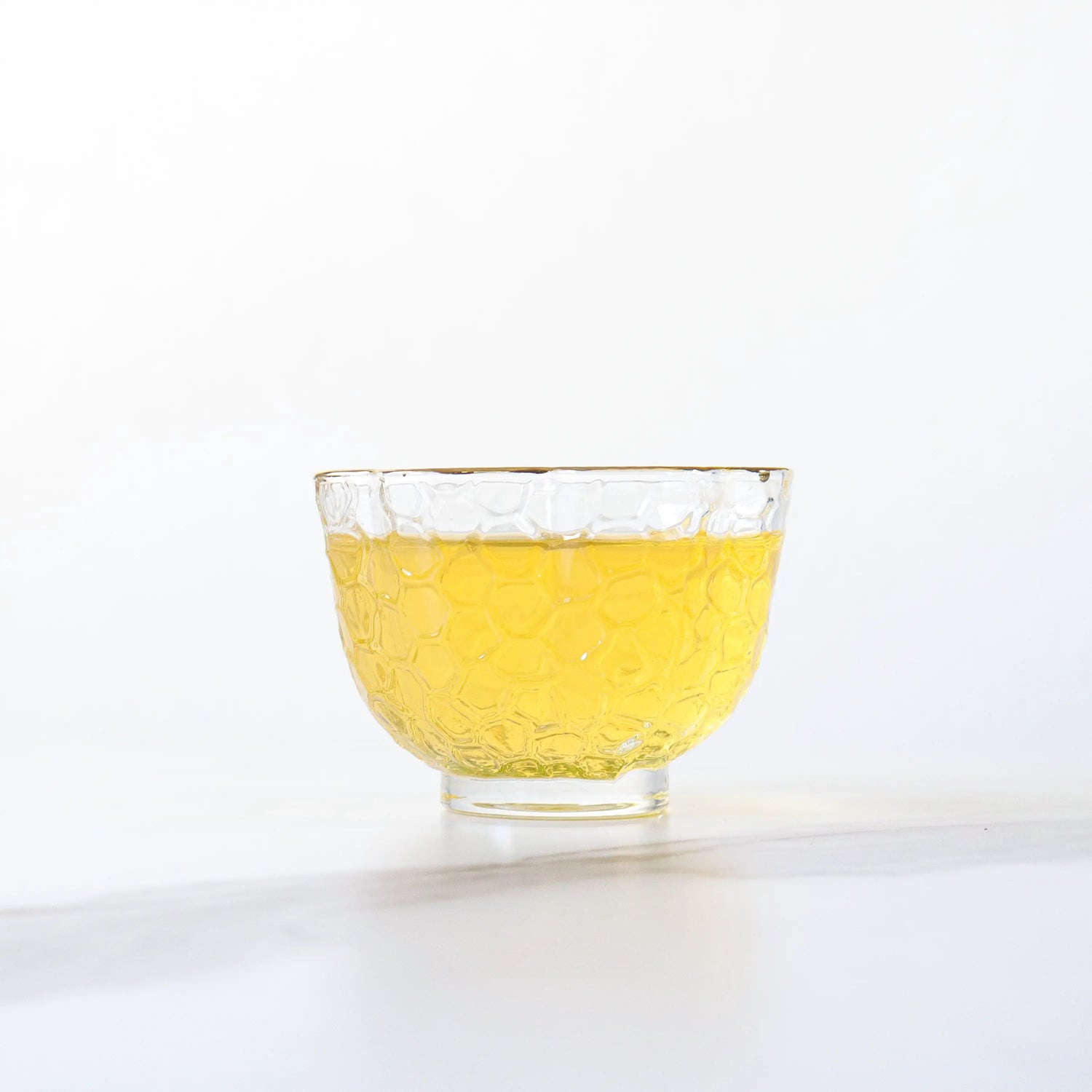 A small, clear glass cup with a textured pattern holds light yellow Jasmine Tea by My Store, sitting on a white surface against a pale background.