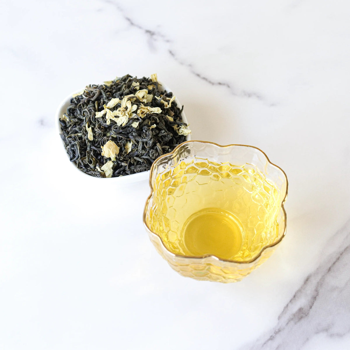 Brewed Jasmine Green Tea beside loose leaves and jasmine petals on white marble.