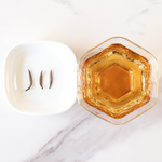 A hexagonal glass of amber liquid is placed beside a white dish with three dried Jin Jun Mei Black Tea leaves from My Store, all set on a white marble surface.