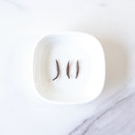 Three dried brown seed pods, arranged on a white square dish, evoke the earthy tones of My Store’s Jin Jun Mei Black Tea—reminiscent of Wuyi Mountain loose leaf black tea—presented on a white marble surface.