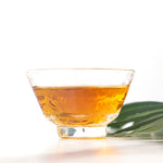 A clear glass cup filled with amber-colored My Store Jin Jun Mei Black Tea loose leaves rests on a white surface beside a green leaf.