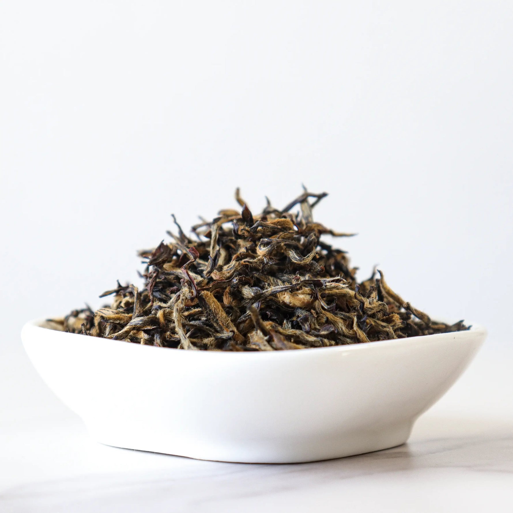 A white bowl of My Store’s Jin Jun Mei Black Tea loose leaves sits on a white surface, highlighting the tea’s Wuyi Mountain heritage against a plain background.