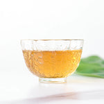 A clear glass cup with a honeycomb pattern holds amber Keemun Black Tea from My Store, placed on a white surface with a green leaf in the background.