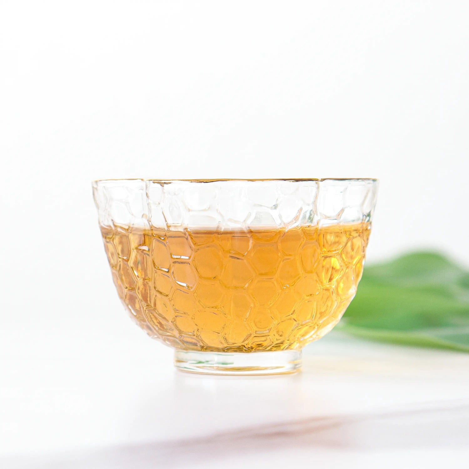 A clear glass cup with a honeycomb pattern holds amber Keemun Black Tea from My Store, placed on a white surface with a green leaf in the background.