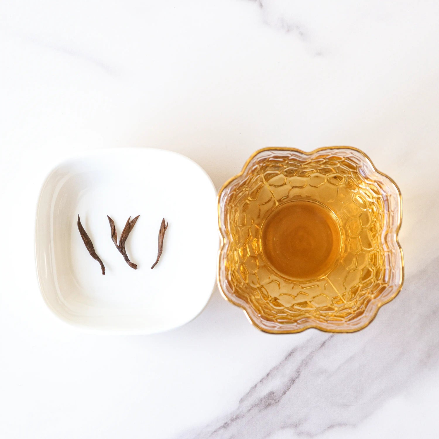 A white bowl with four Keemun Black Tea leaves from My Store sits beside a glass cup of light amber tea on a white marble surface.