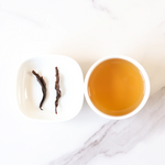 A white dish with two pieces of Lapsang Souchong Black Tea by My Store sits beside a white cup of light amber tea on a marble surface, highlighting the signature smoky aroma of this unique black tea.