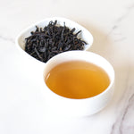 A cup of brewed Lapsang Souchong Black Tea by My Store sits beside a small dish of loose black tea leaves, both on a light surface, inviting you to enjoy their signature smoky aroma.
