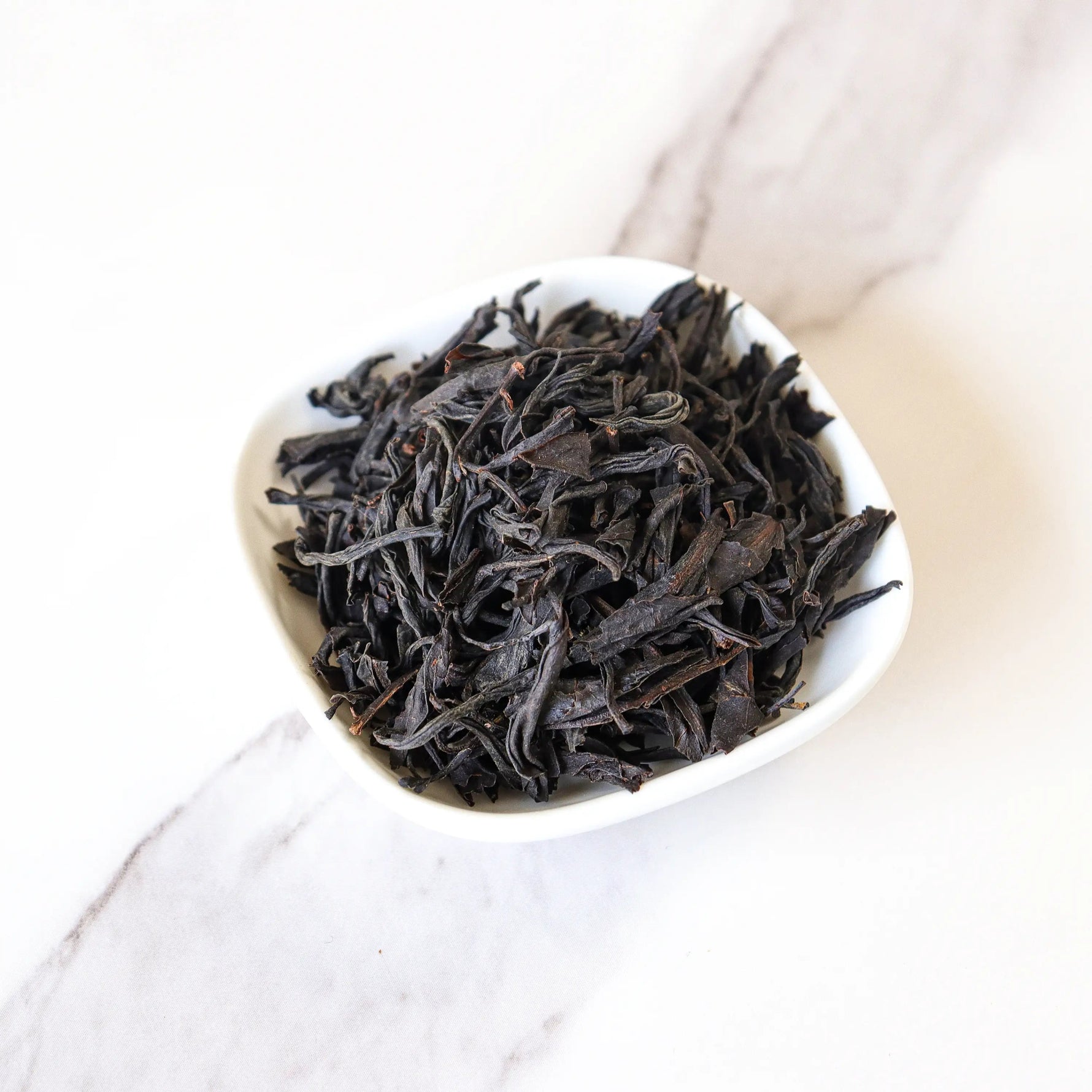 A small white bowl filled with My Store’s Lapsang Souchong Black Tea, renowned for its smoky aroma, rests on a light marble surface.