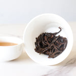 A white cup with brewed My Store Lapsang Souchong Black Tea sits next to a cup of light brown black tea on a white surface, highlighting the tea’s distinctive smoky aroma.