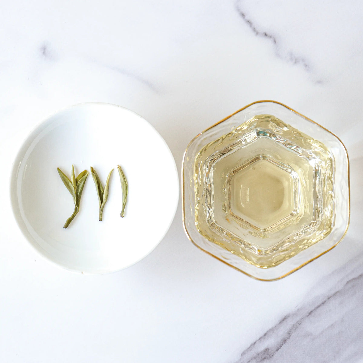 Longjing Dragon Well Green Tea leaves in a tasting dish beside a clear cup of tea.
