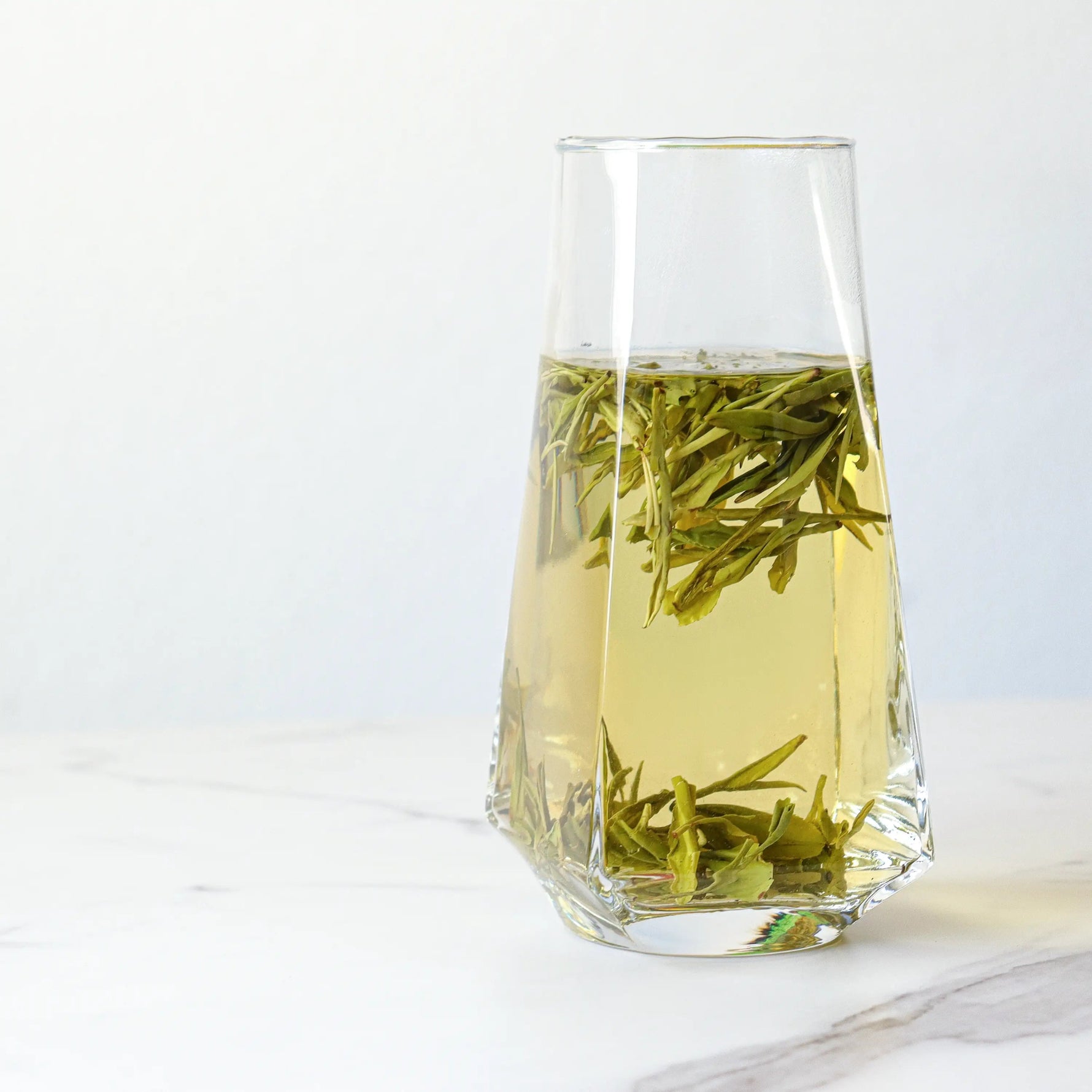 A clear glass of My Store Longjing Dragon Well Green Tea reveals light yellow-green liquid and floating leaves, sitting atop a white marble surface and highlighting its signature chestnut aroma in a minimal setting.