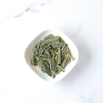 A white bowl of My Store’s Longjing Dragon Well Green Tea, celebrated for its delicate chestnut aroma, rests on a white marble surface.